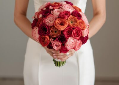 Biedermeier Brautstrauß mit roten, rosa und pfirsichfarbenen Rosen in der Hand einer Braut in einem schlichten weißen Kleid.