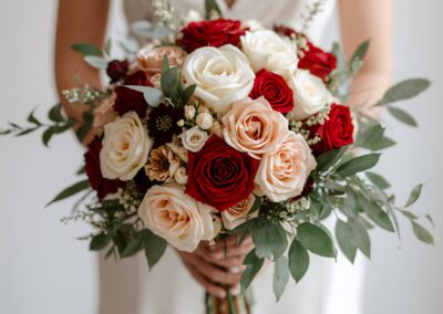 Braut mit einem Strauß aus roten, weißen und roséfarbenen Rosen sowie grünen Blättern in der Hand.