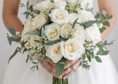 Braut hält einen eleganten Strauß aus weißen Rosen und grünen Akzenten in den Händen.