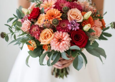 Braut hält einen bunten Blumenstrauß mit roten, rosa und orangefarbenen Blumen in der Hand.