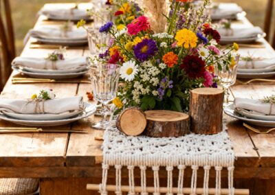 Tischdekoration mit bunten Blumenarrangements und Makramee-Elementen auf einem Holztisch im Freien.